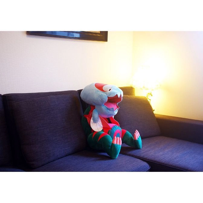  Plush toy of Dracovish sitting on a couch. It has a blue head and green body.