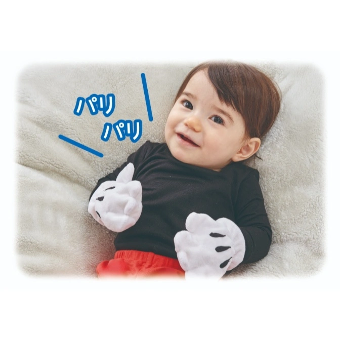  Baby in Mickey Mouse costume with oversized gloves, lying on a blanket and smiling.