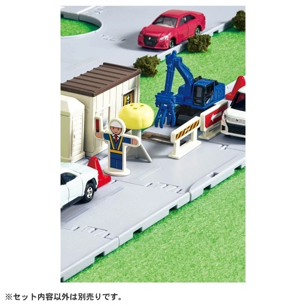 Toy construction site with cars, excavator, building, road worker, and traffic barriers.
