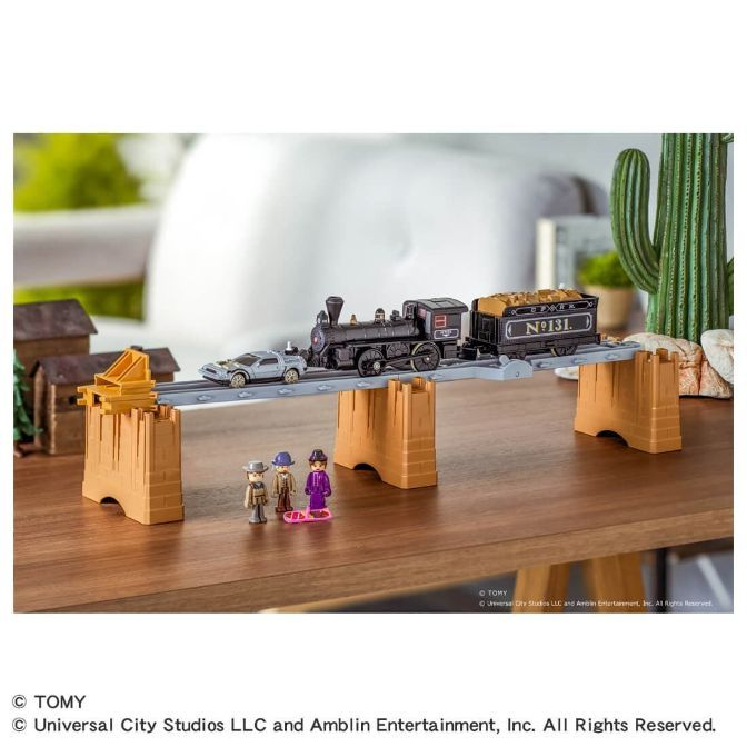  A toy train set with a black steam engine and a silver DeLorean car on a bridge, with three miniature figures standing nearby.