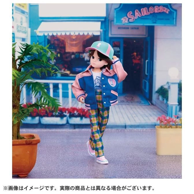  Jenny doll in a varsity jacket, plaid pants, and cap stands in front of a miniature storefront.