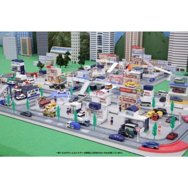  Miniature toy city with buildings, cars, and figures on a green surface.