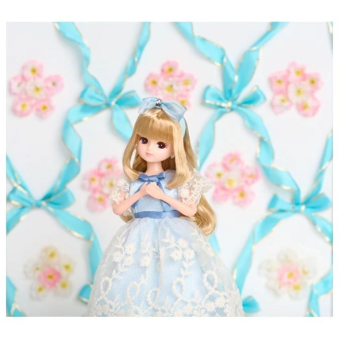  Blonde doll in a blue dress with lace overlay, standing in front of a background of ribbons and flowers.